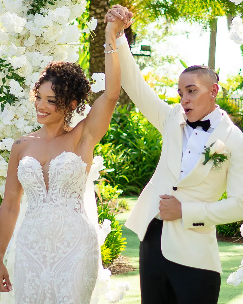 A couple raises their joined hands in celebration beside a white floral arch.