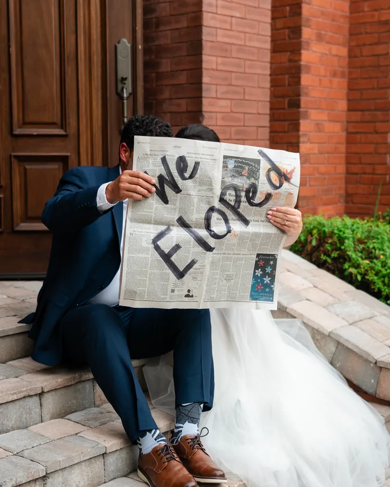 A newlywed couple sits on brick steps behind a newspaper that reads "We Eloped".